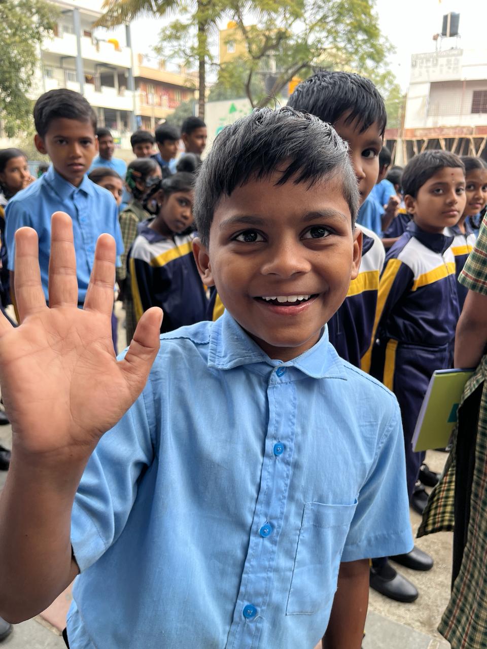A smiling boy in school uniform waves at the camera